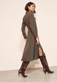 Olivenfarvet langærmet kjole med bælte i taljen, sideslids og knapdetaljer, sammen med blanke bordeaux knæhøje stilethæle.