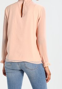 Blouse légère à manches longues de couleur pêche avec une ouverture en forme de goutte dans le dos. Comprend des poignets froncés et une texture subtile de plis verticaux. Portée avec un jean ajusté.