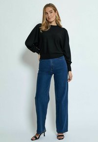 Schwarzer Pullover mit rundem Ausschnitt und langen, lockeren Ärmeln. Kombiniert mit hochgeschnittenen, weiten blauen Jeans und schwarzen Sandalen.
