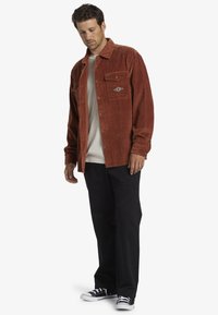 Rust-colored corduroy shirt jacket with two front pockets, a subtle logo, paired with a light shirt and black pants, and black sneakers.