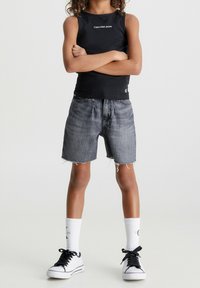 Top negro sin mangas con el logo de "Calvin Klein Jeans", pantalones cortos de mezclilla gris con dobladillo deshilachado, calcetines blancos y zapatillas negras. Un atuendo simple y casual.