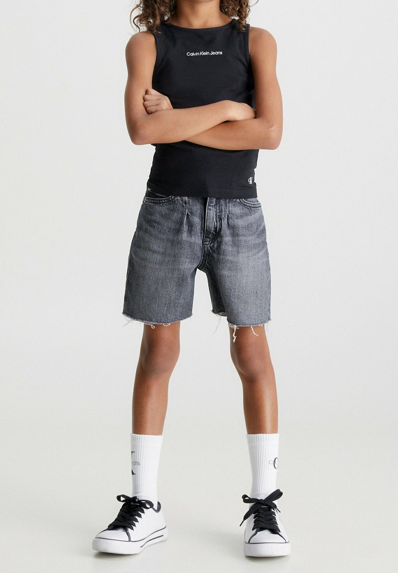 Top negro sin mangas con el logo de "Calvin Klein Jeans", pantalones cortos de mezclilla gris con dobladillo deshilachado, calcetines blancos y zapatillas negras. Un atuendo simple y casual.