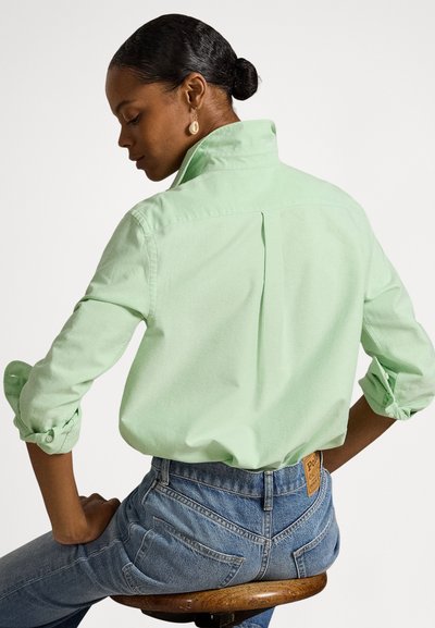 Gaiši zaļa, ar garām piedurknēm krekls ar augstu apkakli un pogātām aprocēm, valkāts kopā ar zilām džinsa biksēm. Raksturīga aizmugurējā šķēluma detaļa.