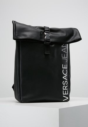 Sac à dos en cuir noir avec un dessus enroulé, sécurisé par une boucle argentée. Présente des lettres verticales blanches sur un côté. Texture lisse, forme structurée.