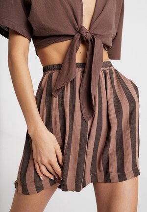 Femme portant un haut court marron noué à la taille et un short taille haute à rayures verticales marron et rose, main posée sur la cuisse.