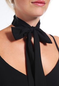 Black fabric neck bow, featuring two long tails and a smooth texture, tied in a large bow at the front, attached to a sleeveless garment.