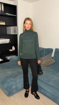 Green turtleneck top paired with black flared pants and black loafers. Hair down, standing against a textured teal couch with cushions.
