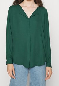 Blusa di tessuto verde con scollatura a V e maniche lunghe, caratterizzata da una leggera lucentezza e una consistenza morbida; cade sotto la vita, abbinata a jeans in denim chiari.