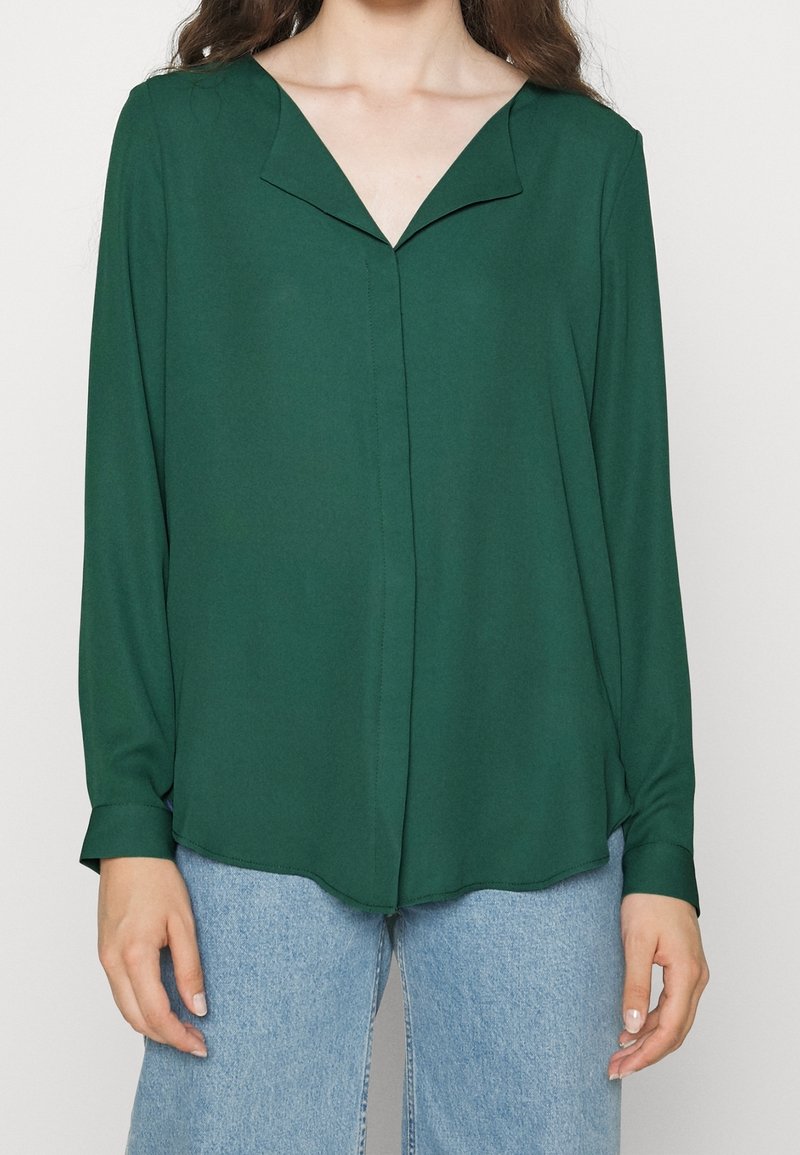 Blusa di tessuto verde con scollatura a V e maniche lunghe, caratterizzata da una leggera lucentezza e una consistenza morbida; cade sotto la vita, abbinata a jeans in denim chiari.