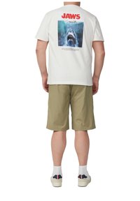 Witte t-shirt met een haai-afbeelding op de achterkant en "JAWS" in vet rood lettertype. Draagt bij groene shorts en witte sneakers.