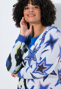Kleurvolle cardigan van een witte gebreide stof, versierd met zwarte, blauwe, roze en gele sterpatronen. Het heeft een V-hals en geribbelde manchetten.