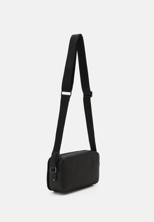 Petit sac bandoulière en cuir noir compact avec une sangle large réglable et des détails de boucle argentés sur un fond uni.