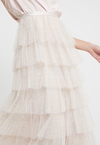 Jupe en tulle à volants superposés couleur beige clair, avec plusieurs couches froncées ornées de motifs étoiles subtils et une ceinture en satin. Texture douce.