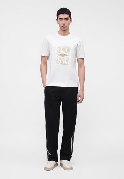 Νέος άνδρας που φοράει λευκό T-shirt Emporio Armani, μαύρο παντελόνι με λευκές ρίγες και μπεζ αθλητικά, στέκεται μπροστά σε απλό φόντο.