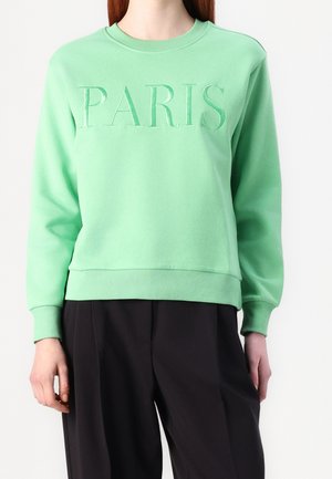 Vrouw draagt een mintgroene sweater met "PARIS" in reliëf op de borst, gecombineerd met losse zwarte broek, tegen een witte achtergrond.