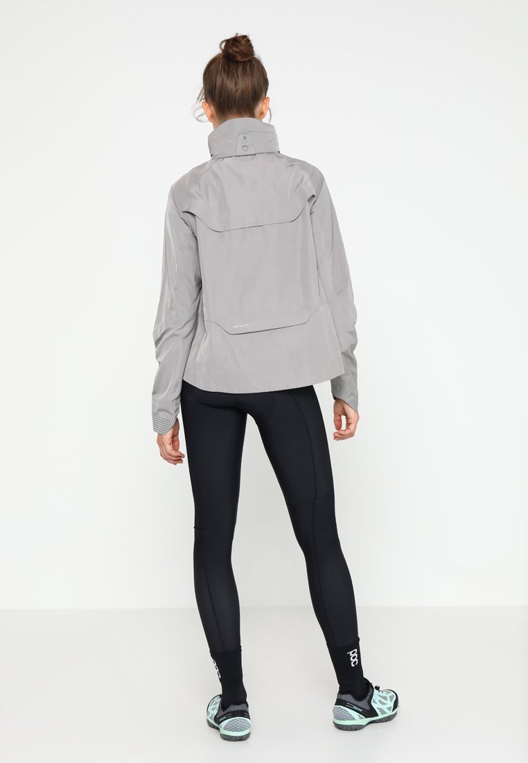 Hellgraue wasserdichte Jacke mit hohem Kragen und Rückentasche, kombiniert mit schwarzen Leggings und türkisfarbenen Schuhen. Glatte Textur, sportliches Design.