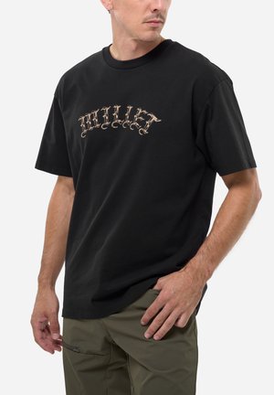 Homme portant un T-shirt noir avec "MILLET" en lettres gothiques sur la poitrine et un pantalon vert olive, debout avec une main dans la poche.