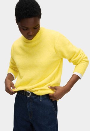 Gul strikket sweater med ribbet rund hals og lange ærmer, kombineret med mørkeblå jeans. Sweateren har en blød tekstur og kontrastfarvede manchetter.