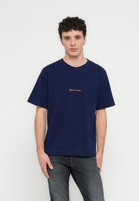 T-shirt in cotone blu navy con maniche corte, scollo rotondo e piccolo dettaglio di testo arancione "Original Studio" sul petto. Modello con jeans grigi.