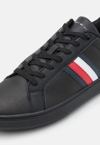 Musta nahkainen lenkkari, jossa on teksturoitu pinta ja harmaat, valkoiset sekä punaiset raidat sivulla. Perinteinen nauhakiinnitys.