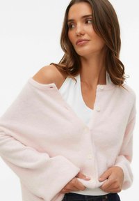 Gaiši rozā, pārlielots kardigans no mīkstas auduma. Tam ir pogu aizdare, ribota aproce un atvērts dizains, atklājot baltu krekliņu zem tā.