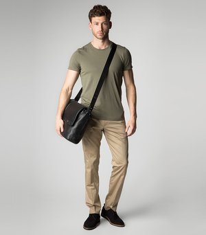 Borsa a tracolla in pelle nera con pattina, indossata a croce; t-shirt aderente verde oliva, pantaloni beige e scarpe nere. Design semplice.