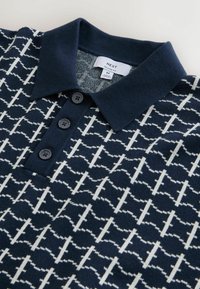 Marineblauw poloshirt met wit geometrisch patroon, drie zwarte knopen en een effen kraag. Gemaakt van zachte stof, met een label dat een ontspannen pasvorm aangeeft.
