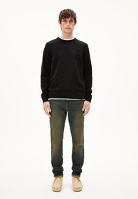 Schwarzer Pullover mit strukturiertem Finish, weißem Kragenbesatz und Logo auf der Brust, kombiniert mit verblassten grünen Jeans und beigen Schuhen mit weicher Sohle.