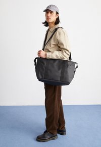 Schwarze Tragetasche mit dunklen Trägern und einer strukturierten Oberfläche, ausgestattet mit einem ausgeprägten Logo und einem geräumigen Design, die über der Schulter getragen wird.