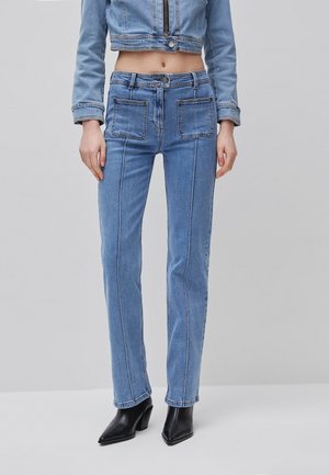 Frau trägt hoch taillierte blaue Jeans mit aufgesetzten Taschen vorne, eine kurze Jeansjacke und schwarze spitze Stiefeletten.