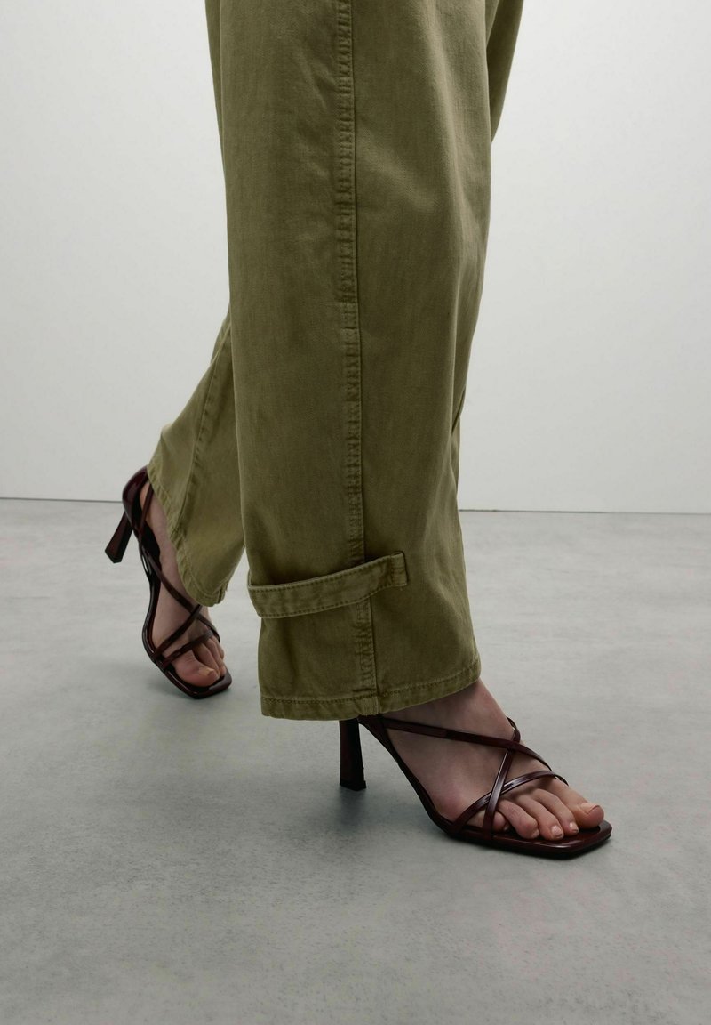 Strappy brown high-heeled sandals with a square toe; paired with loose, olive green, cuffed trousers. Smooth texture, minimal detailing.