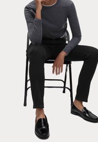 Grå långärmad skjorta, svarta slim-fit byxor och glänsande svarta loafers, sittande på en svart hopfällbar stol mot en vit bakgrund.