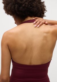 A peça de roupa burgundy com decote halter apresenta uma silhueta ajustada, textura suave e detalhes nas costas abertas, destacando o ombro e a coluna.