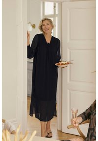 Black, sheer maxi dress with long sleeves, gathered detail, and a tied neckline. The model holds a plate of food in a bright, interior setting.