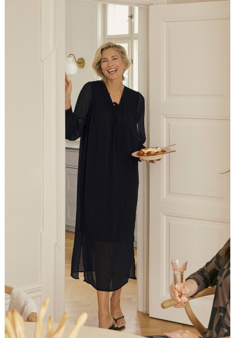 Black, sheer maxi dress with long sleeves, gathered detail, and a tied neckline. The model holds a plate of food in a bright, interior setting.