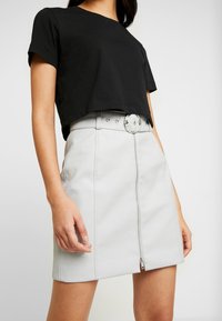 Zwarte cropped t-shirt gecombineerd met een lichtgrijze faux leren rok met een gestructureerd oppervlak en een rits aan de voorkant, geaccentueerd met een ronde riemgesp.
