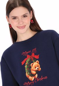 Tengerészkék pulóver, amelyen egy medve látható, amint egy karácsonyi koszorút tart. Piros szöveggel, "Boldog Karácsonyt" felirattal és díszes szalagokkal díszített grafikai mintával.
