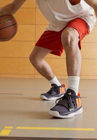 Black athletic shoes with orange stripes and white soles, worn with red shorts and a white mesh tank top on a basketball court.
