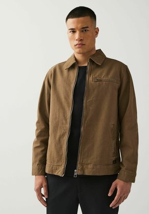 Brown canvas jacket with a collar, front zipper, and a chest pocket. Worn over a black shirt, featuring a slightly textured finish.