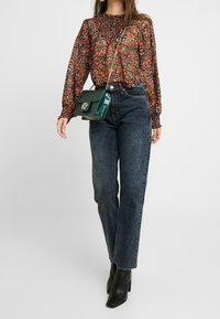 Blusa nera trasparente a fiori con scollatura arricciata, abbinata a jeans blu scuro a vita alta e stivaletti neri con tacco; borsa a tracolla verde.