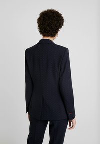Blazer bleu marine à pois blancs et rouges, coupe ajustée, tissu texturé, et boutons d'accent rouges sur les poignets. Vue de dos.