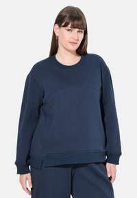 Donna con capelli castani lisci, che indossa una felpa a maniche lunghe blu navy e pantaloni coordinati, in piedi davanti a uno sfondo semplice.