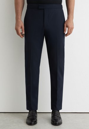 Mies, jolla on räätälöidyt tummansiniset juhlahousut ja mustat nahkaiset loaferit, seisoo vaaleaa seinää ja lattiaa vasten.