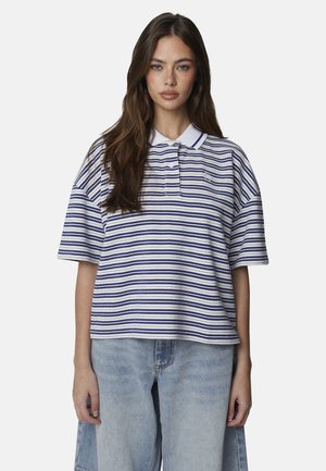 Giovane donna con lunghi capelli castani che indossa una polo a righe bianche e blu e jeans azzurri, in piedi contro uno sfondo semplice.