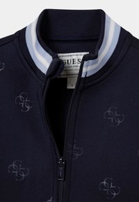 Blouson aviateur bleu marine doté d'une fermeture éclair et d'un col côtelé à rayures bleues et blanches. Motif logo subtil sur toute la surface du tissu.