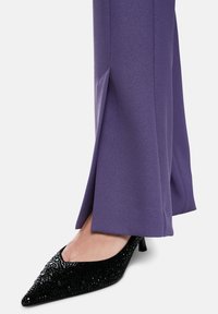 Purple, tailored trousers with a subtle sheen, featuring a split detail at the hem, paired with black, embellished, pointed-toe heels.