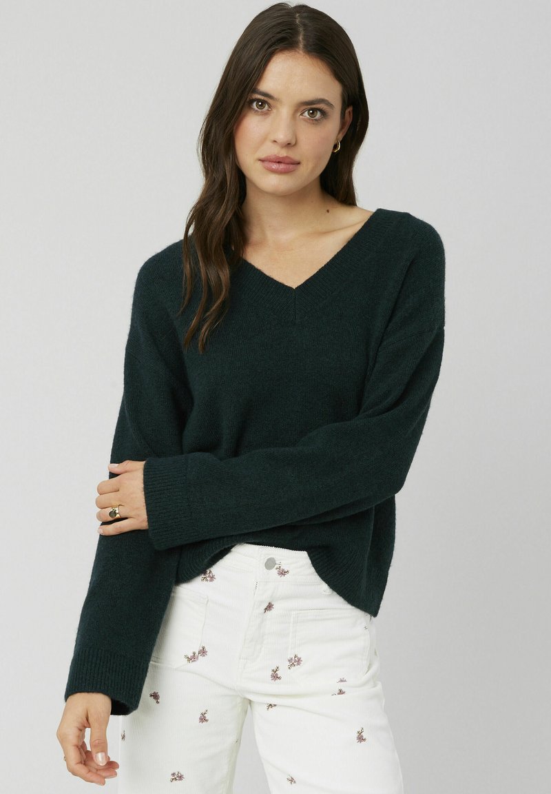 Pull vert foncé en V à manches longues, fabriqué dans un matériau doux. Présente des poignets côtelés et une coupe décontractée, associé à un pantalon brodé blanc.