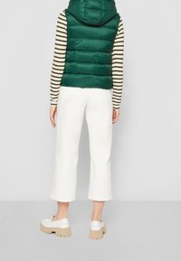 Zielona pikowana kamizelka z kapturem noszona na paskowanej, długim rękawie koszuli. Dobrana z białymi spodniami o szerokich nogawkach i beżowymi, platformowymi butami wsuwanymi.