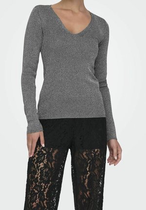 Žena má na sobě šedý přiléhavý žebrovaný top s dlouhým rukávem a černé průsvitné kalhoty s květinovou krajkou a černou podšívkou.