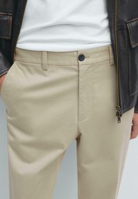 Chinos beige avec une texture lisse, dotés d'une poche avant, d'un bouton noir de fermeture et d'une discrète fermeture éclair sur le côté.